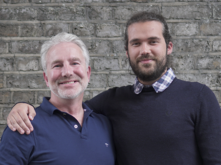 Two men (Ben Shoulder and Ben Graham) standing side by side in front of a brick wall, smiling at the camera, one older with grey hair in a navy polo shirt and the other younger with dark hair in a bun wearing a navy sweater.
